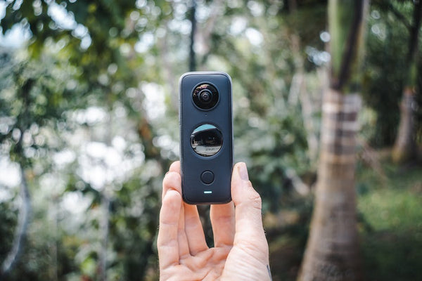 Close-up of a hand holding a compact 360 camera outdoors with greenery.