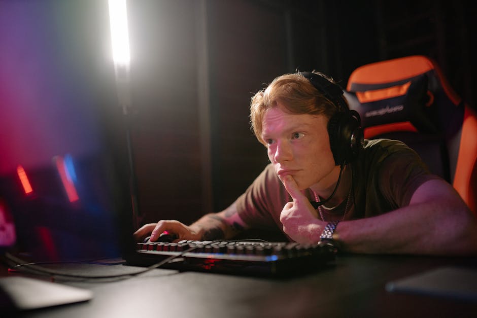 A young man intensely gaming on a computer, surrounded by tech gear in a dimly lit room.
