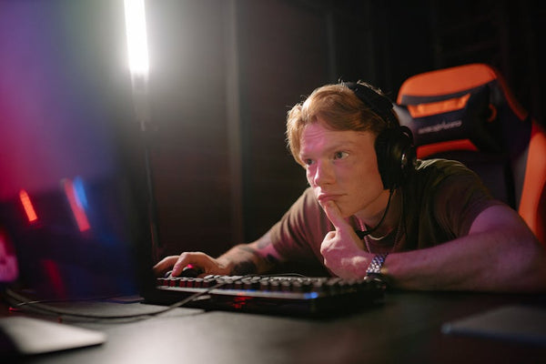 A young man intensely gaming on a computer, surrounded by tech gear in a dimly lit room.